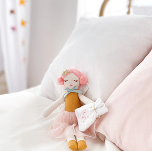 The MON AMI Tooth Fairy Doll with Pouch by Mon Ami, featuring pink hair and a pink dress, sits on a bed next to two pillows, guarding its small tooth pouch in anticipation of the Tooth Fairy.