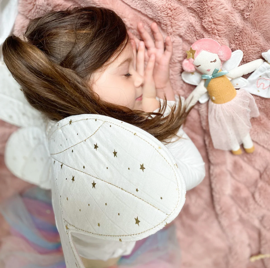 A child with white, star-patterned fairy wings drifts off to sleep on a pink furry blanket beside the MON AMI Tooth Fairy Doll with Pouch by Mon Ami, which dons a pink hat and fairy wings. The child dreams of the Tooth Fairy arriving soon to gather the tiny treasure securely stored in their magical tooth pouch.