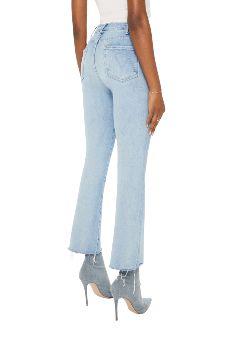 A person wearing light blue, high-rise flare jeans with a raw hem and gray high-heeled shoes is standing with their back to the camera. They are also wearing a white top from Mother Denim The Hustler Ankle Fray by Mother.