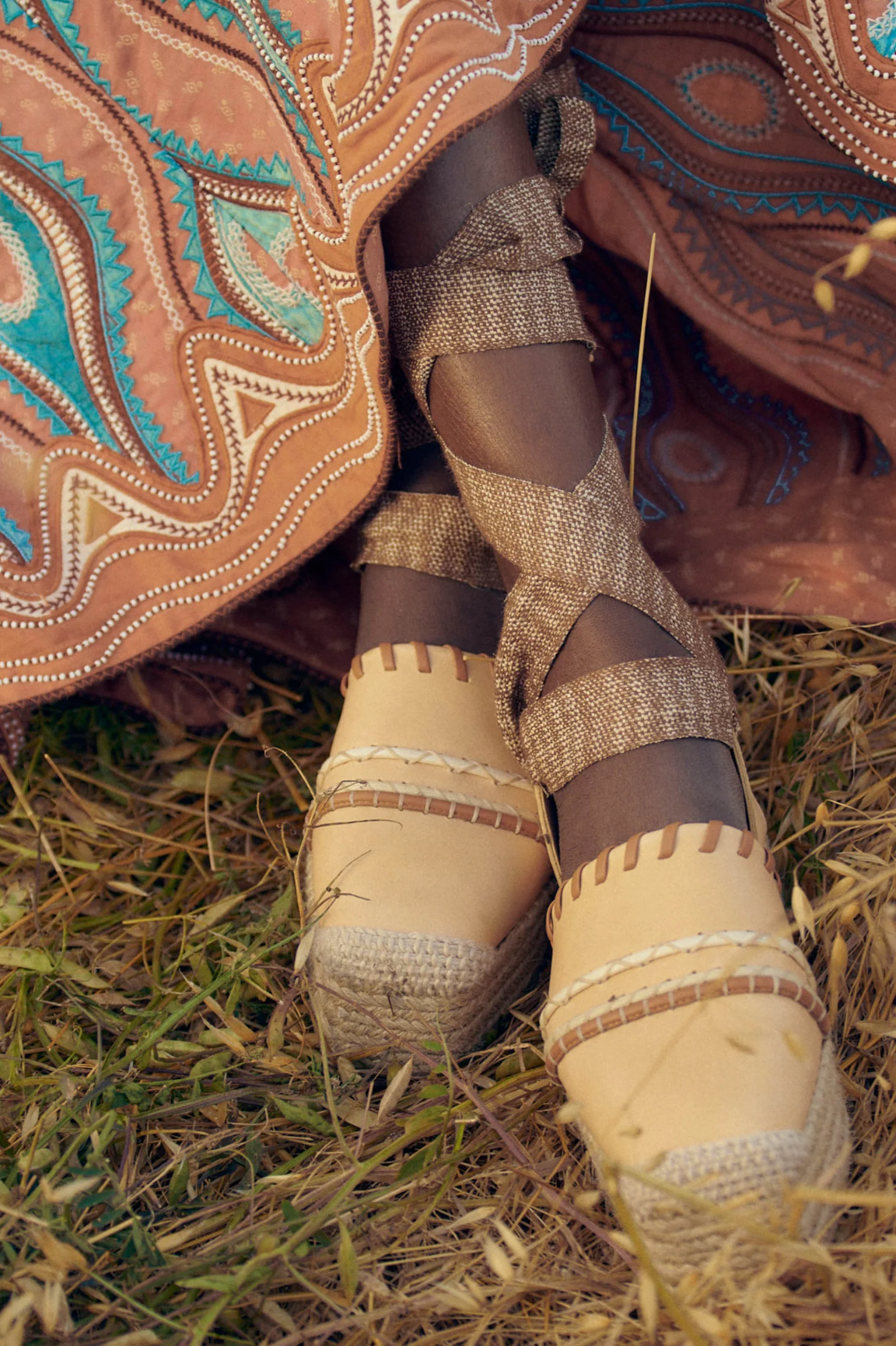 Close-up of a person's feet wearing Ulla Johnson Concha Platform Espadrille shoes with lace-up straps, standing on grass. The person is wearing a colorful, patterned dress.