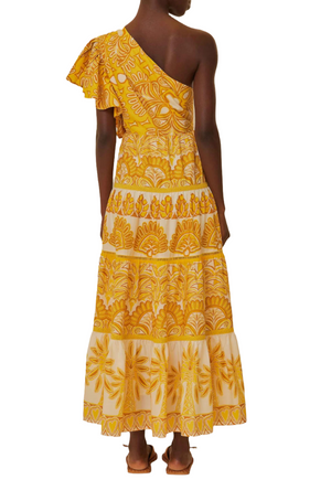 A person stands facing away, wearing the stunning Farm Rio Ainika Tapestry Yellow One Shoulder Maxi Dress with a tiered skirt in yellow and white floral print, complemented by flat sandals.