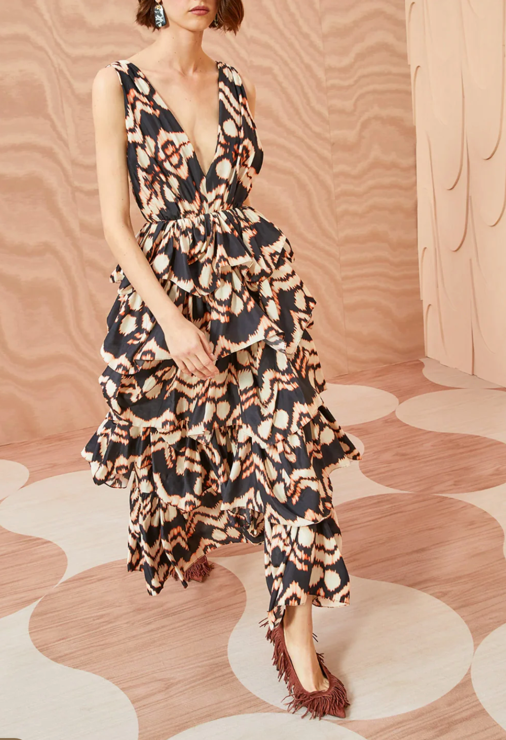 A person is walking on a patterned floor wearing the Ulla Johnson Dephne Dress, a sleeveless, v-neck, tiered ruffle midi dress adorned with an abstract design in black, white, and beige. The background features a matching abstract pattern. They stand out in their Ulla Johnson dress as the ruffles of the midi skirt blend harmoniously with the intricate floor design.