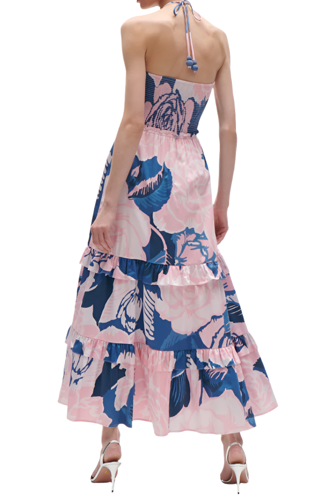 A person is wearing the Figue June Dress, a halter-neck, sleeveless piece adorned with a layered floral pattern and high heels. The dress showcases pink and blue hues and features an adjustable sweetheart neckline. The person is photographed from the back.