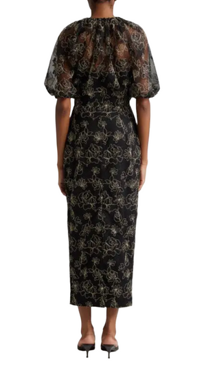 The individual stands facing away, wearing the Lela Rose Naomi Dress, a black designer sheath dress adorned with floral patterns and billowed sheer puff sleeves by Lela Rose.