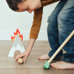 A child in a brown shirt and jeans plays with the wooden balls and toy mallet from the Asweets Putt Putt Golf Set by Asweets, near a small, toy campfire structure, reminiscent of a quaint golf setup.