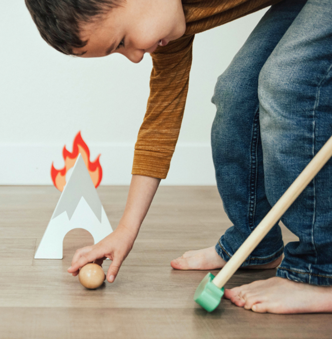 A child in a brown shirt and jeans plays with the wooden balls and toy mallet from the Asweets Putt Putt Golf Set by Asweets, near a small, toy campfire structure, reminiscent of a quaint golf setup.