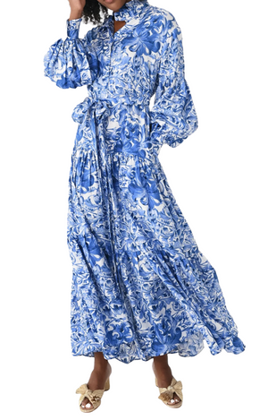 Person wearing the Evi Grintela Elsa Silk Dress, a long maxi shirtdress featuring a blue and white floral print, puffed sleeves, and a tied waist, paired with sandals adorned with bows.