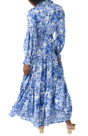 A person with curly hair wearing the Evi Grintela Elsa Silk Dress, a long blue and white floral print maxi shirtdress, and beige heeled sandals is seen from the back.