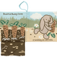 The Jellycat Bashful Bunny Loves You Book, crafted by Jellycat, features a baby-friendly, soft cloth page where Bashful Bunny is depicted holding a carrot alongside the text "Bashful Bunny loves crunching on carrots." The illustration also shows carrots growing in the ground.