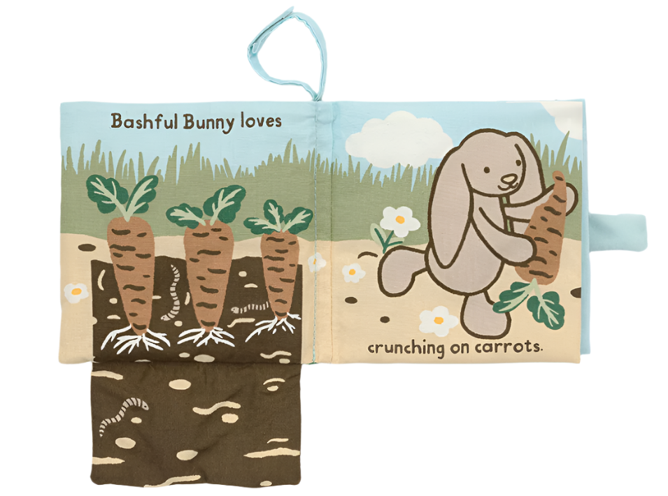 The Jellycat Bashful Bunny Loves You Book, crafted by Jellycat, features a baby-friendly, soft cloth page where Bashful Bunny is depicted holding a carrot alongside the text "Bashful Bunny loves crunching on carrots." The illustration also shows carrots growing in the ground.