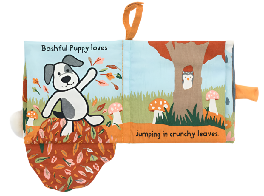 A page from the Jellycat Bashful Puppy Loves You Book features an adorable dog amidst leaves with the text: "Bashful Puppy loves jumping in crunchy leaves." The charming background of this delightful cloth baby book by Jellycat showcases a tree adorned with mushrooms and an owl nestled in a hole.