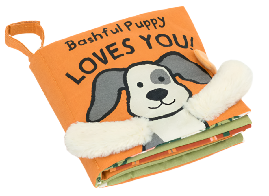 The Jellycat Bashful Puppy Loves You Book by Jellycat features an orange cloth cover adorned with a cartoon dog and the text "Bashful Puppy Loves You!" The book includes charming details such as fuzzy ears that stick out and green pages inside.