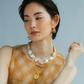 A person with short dark hair wears a sleeveless checked top, a chunky pearl necklace with a 24-karat gold-plated pendant, and Lizzie Fortunato Onda Hoops by Lizzie Fortunato, gazing to the side.