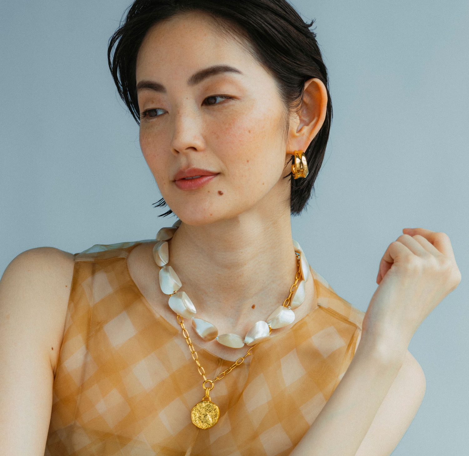 A person with short dark hair wears a sleeveless checked top, a chunky pearl necklace with a 24-karat gold-plated pendant, and Lizzie Fortunato Onda Hoops by Lizzie Fortunato, gazing to the side.