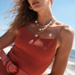 A person in a textured rust-colored dress with layered necklaces stands on a beach, accentuating their look with the Lizzie Fortunato Louise Suede Belt in coral, featuring an oversized buckle.