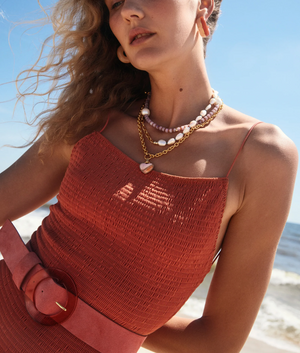 A person in a textured rust-colored dress with layered necklaces stands on a beach, accentuating their look with the Lizzie Fortunato Louise Suede Belt in coral, featuring an oversized buckle.