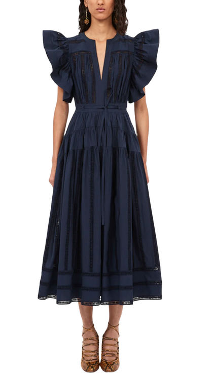 A person wearing the Ulla Johnson Emery Dress, a navy blue creation featuring ruffled shoulders and lace details with geometric eyelet stripes, paired with brown high-heeled shoes.