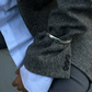 Close-up of a person wearing a grey herringbone blazer with silver cuff over a white shirt and grey trousers. The left sleeve features three buttons, with the hand positioned near the side, showcasing a LIÉ STUDIO Amanda Bracelet made from gold-plated brass by Lie Studio.