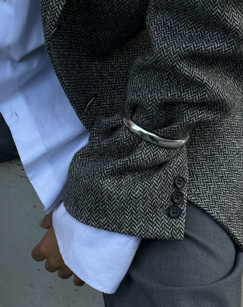 Close-up of a person wearing a grey herringbone blazer with silver cuff over a white shirt and grey trousers. The left sleeve features three buttons, with the hand positioned near the side, showcasing a LIÉ STUDIO Amanda Bracelet made from gold-plated brass by Lie Studio.