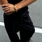 A person standing on a road, wearing black pants and a black top with arms crossed, showcases an exquisite LIÉ STUDIO Amanda Bracelet of gold-plated brass by Lie Studio on one wrist.