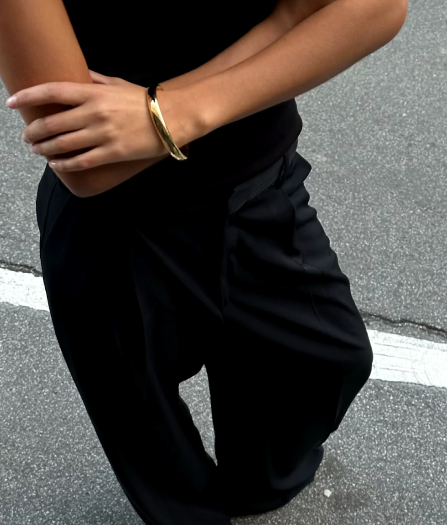 A person standing on a road, wearing black pants and a black top with arms crossed, showcases an exquisite LIÉ STUDIO Amanda Bracelet of gold-plated brass by Lie Studio on one wrist.