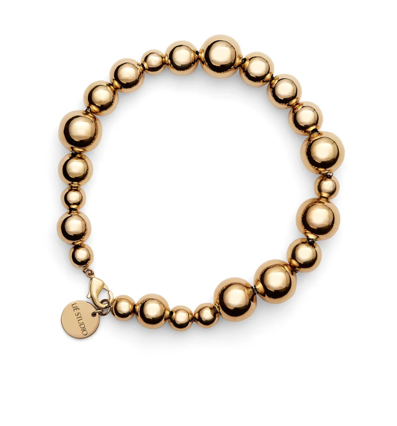 The LIÉ STUDIO Elly Bracelet by Lie Studio features an elegant arrangement of gold round beads in various sizes, complemented by a circular charm with intricate engraving, all showcased beautifully against a white background. This refined piece of pearl jewelry radiates timeless sophistication.