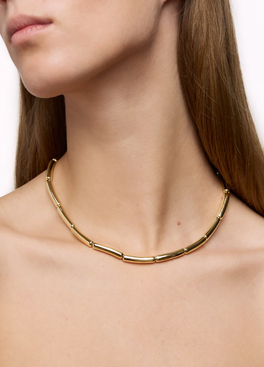 A close-up of a person wearing the elegant LIÉ STUDIO Margaret Necklace by Lie Studio, featuring elongated, tubular gold-plated brass links. The person's face is partially visible, and they have long, brown hair.