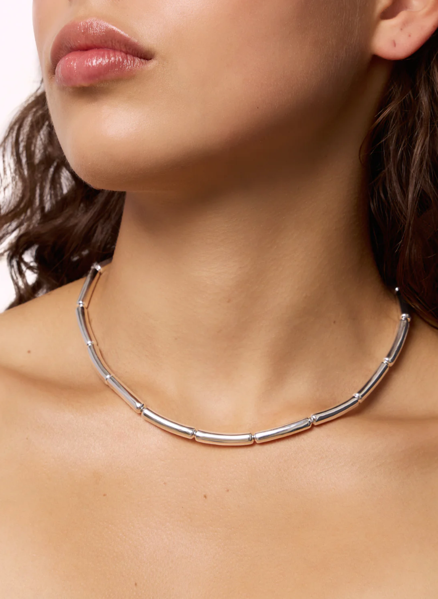 Close-up of a person wearing the LIÉ STUDIO Margaret Necklace by Lie Studio, which features an elegant tubular silver design around their neck. The person has bare shoulders and is framed from the lower face to the upper chest.