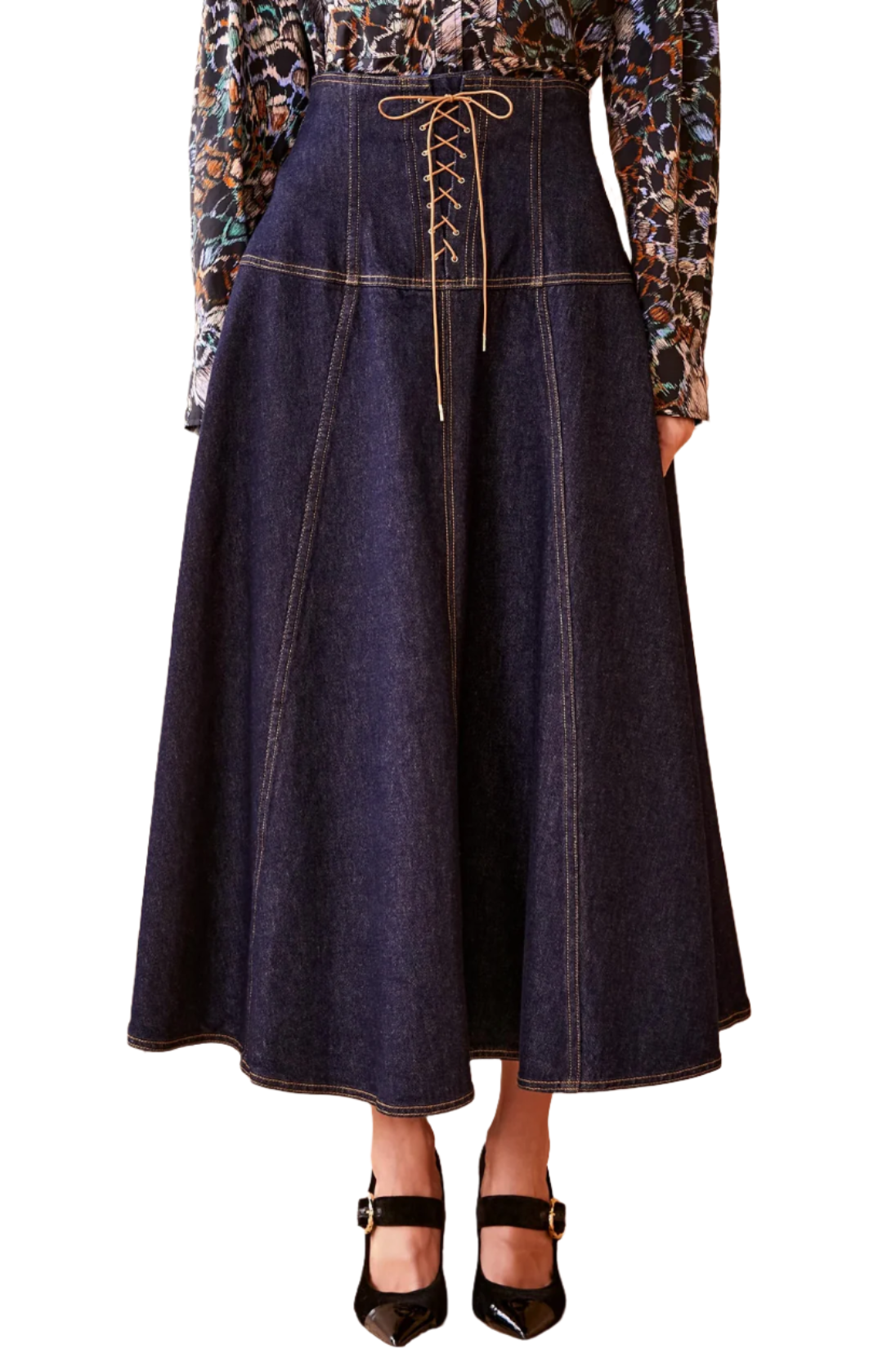 A person wearing the Ulla Johnson The Estelle Denim Skirt in navy blue, featuring a high-waisted A-line silhouette with feminine lace-up detailing in the front, paired with a patterned blouse and black ankle-strap heels.