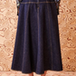 A person is facing a patterned wooden wall, wearing the Ulla Johnson The Estelle Denim Skirt from Ulla Johnson and black shoes with bows at the back. Only the back of the person is visible, highlighting their elegant denim silhouette.