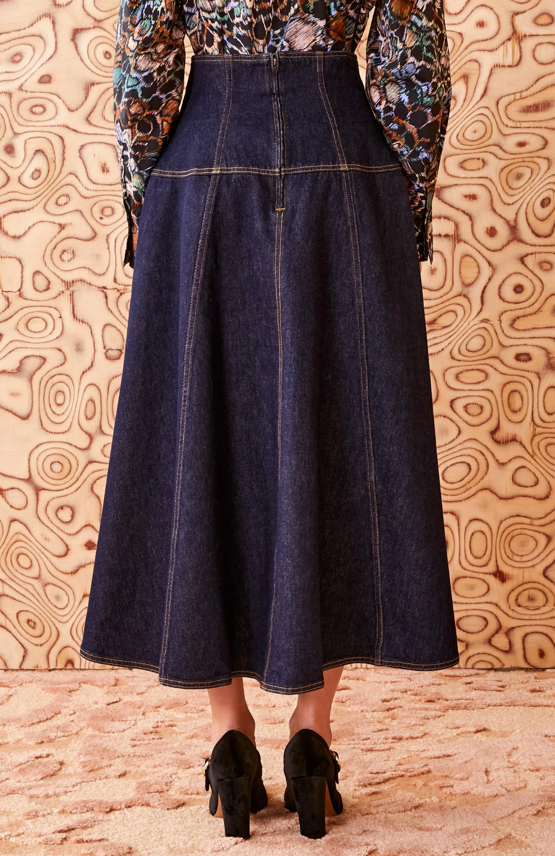 A person is facing a patterned wooden wall, wearing the Ulla Johnson The Estelle Denim Skirt from Ulla Johnson and black shoes with bows at the back. Only the back of the person is visible, highlighting their elegant denim silhouette.