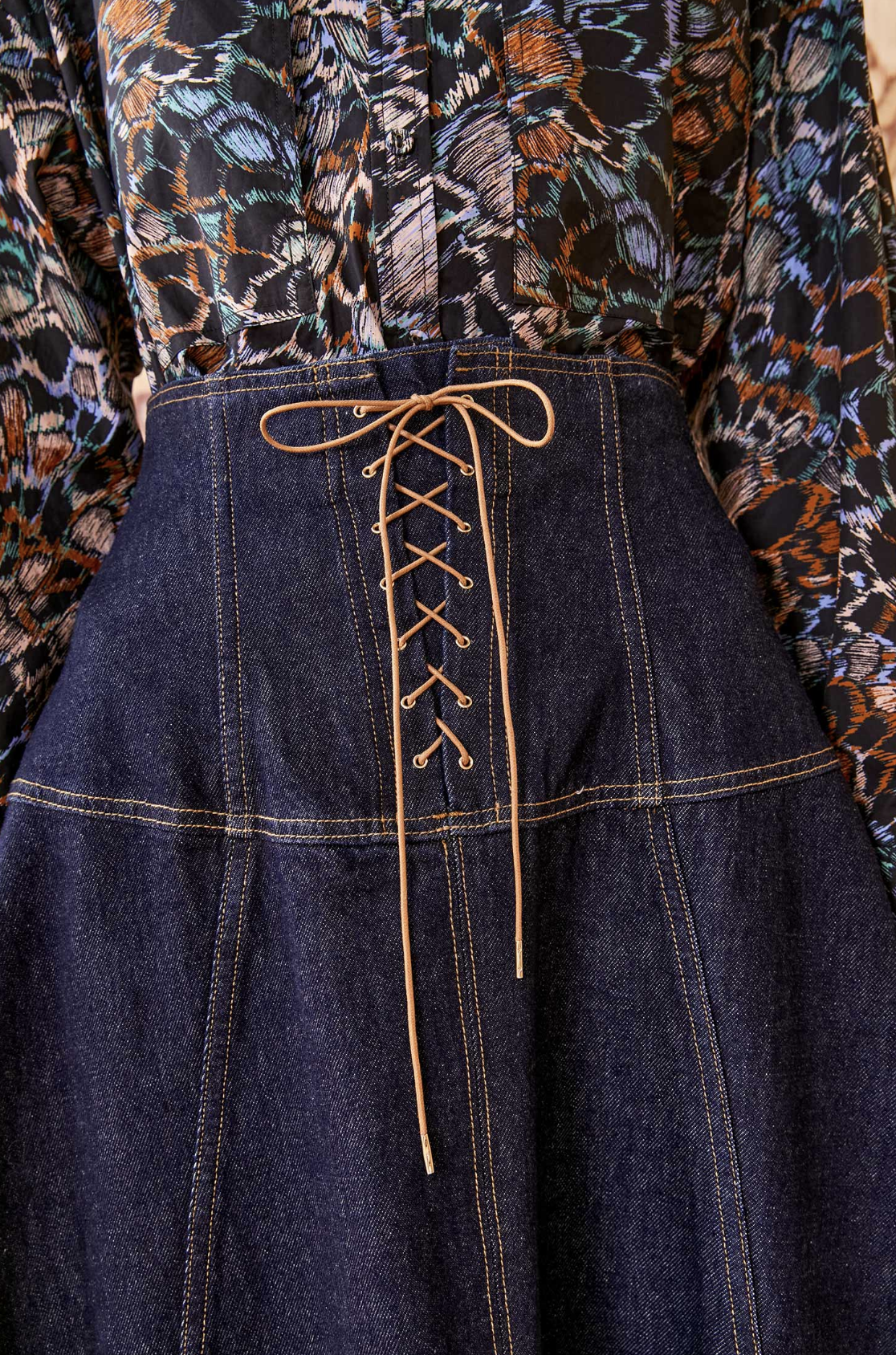 Close-up of a person wearing the Ulla Johnson The Estelle Denim Skirt with feminine lace-up detailing down the front center, paired with a patterned blouse in shades of blue, brown, and black from Ulla Johnson.