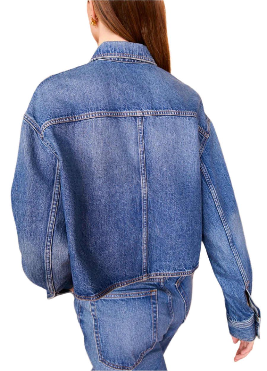 A person in an Ulla Johnson The Bobbi Denim Jacket made of rigid denim and jeans stands with their back to the camera, facing a decorative, textured wall.