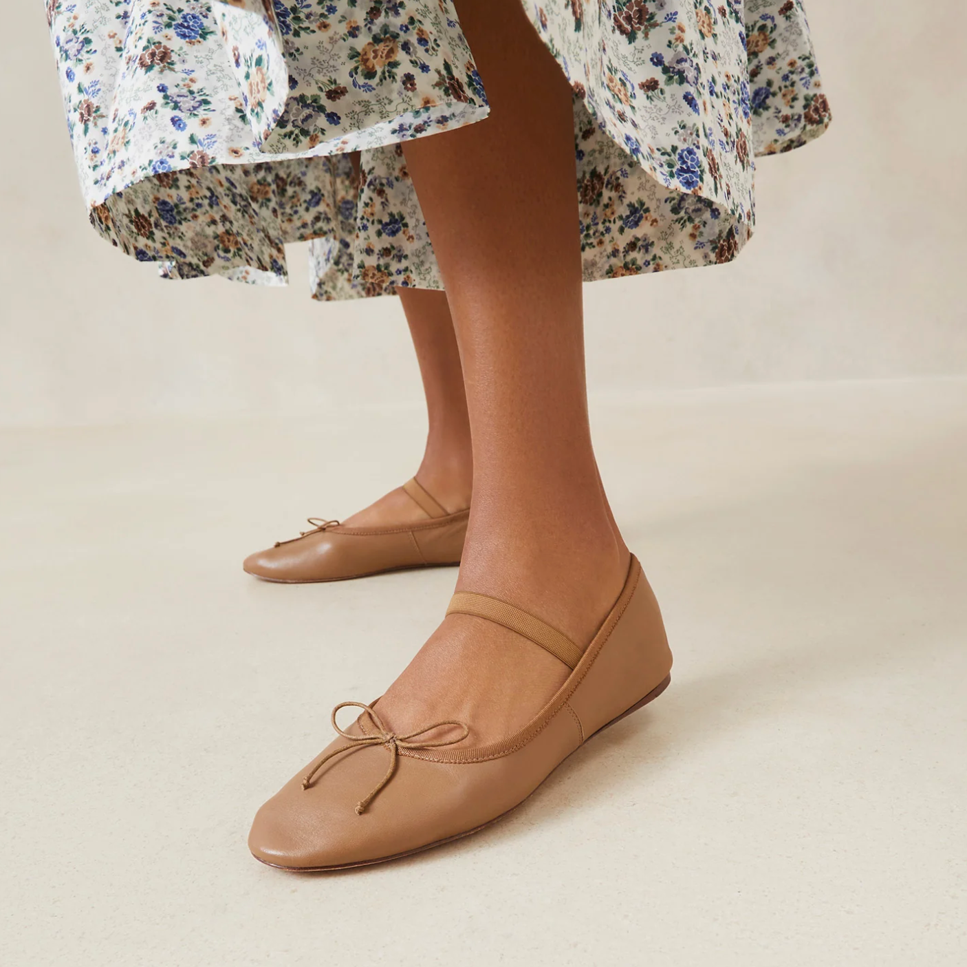 A person wearing a floral dress and Loeffler Randall's Leonie Soft Ballet Flats in tan, featuring small bows and a padded footbed, stands on a smooth beige surface.