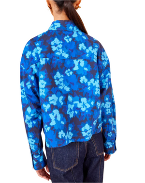 A person wearing an Ulla Johnson Aria Blouse in a vivid blue hue, paired with long sleeves and dark jeans, adorned with eye-catching ikat floral patterns, viewed from the back.