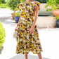 Wearing the Ulla Johnson Rumi Top, featuring a striking yellow and brown floral design inspired by ikat patterns, a person smiles while standing outdoors near a fountain and potted plants.