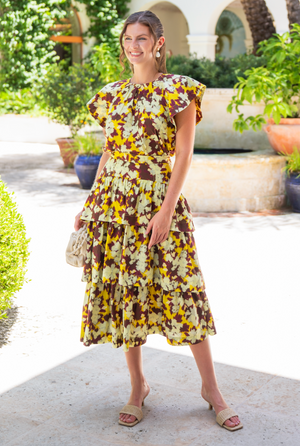 Wearing the Ulla Johnson Rumi Top, featuring a striking yellow and brown floral design inspired by ikat patterns, a person smiles while standing outdoors near a fountain and potted plants.