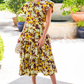 A woman in an Ulla Johnson Winnie Skirt, featuring a floral design and ruffled tiers, stands outdoors on a sunny day, holding a small bag. Behind her, potted plants frame the scene alongside a gently flowing fountain.