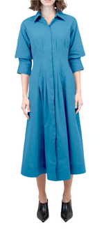 A person in a Simkhai Signature Jazz Dress, a blue long-sleeved midi featuring elegant pintuck detailing and a tailored bodice, stands with arms relaxed. Black pointed shoes are visible against the white background.