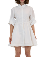 A person wearing the Simkhai Jazzie Mini Dress, a white knee-length button-up dress crafted from crisp poplin with rolled-up sleeves.