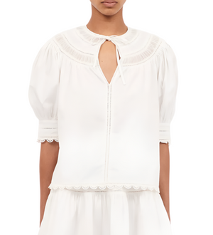 An individual is dressed in the Ulla Johnson Dylan Top, characterized by its white blouse design with a lace inset, puff sleeves, and a tied neckline.