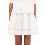 A person wearing a short Ulla Johnson Ayla Skirt in white, featuring a scalloped hem and lace trim, with their hands at their sides.