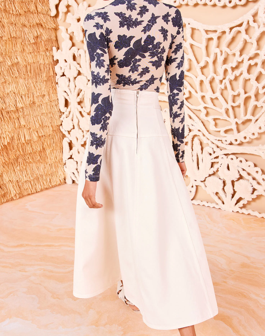 A person wearing the Ulla Johnson Brigette Skirt, a white high-waisted A-line design, paired with a blue floral long-sleeve top, stands elegantly in front of a decorative textured wall.