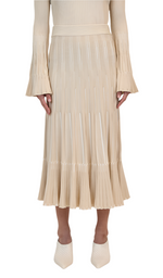 A person in a beige pleated Simkhai Emerie Midi Skirt with long sleeves and white heeled shoes, featuring an A-line silhouette, stands against a plain white background.