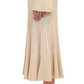 Side view of a person in a beige pleated Simkhai Emerie Midi Skirt with bell sleeves and elegant A-line silhouette, paired with white heeled mules, shown from shoulder to ankle.