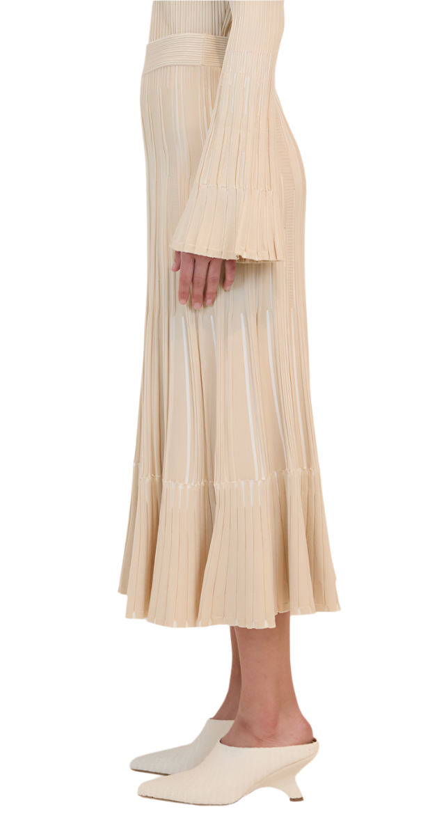 Side view of a person in a beige pleated Simkhai Emerie Midi Skirt with bell sleeves and elegant A-line silhouette, paired with white heeled mules, shown from shoulder to ankle.