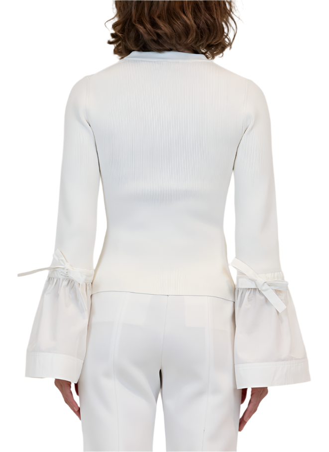 A person showcases elegance in a Simkhai Agata Top, a white ribbed blouse with flared sleeves and tie details, paired with matching white pants.