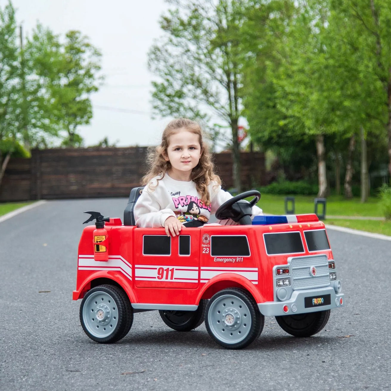 A young girl rides the Freddo Fire Truck on a paved road, surrounded by green trees and a wooden fence, feeling like a true hero.