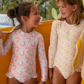 Two young girls in Minnow Girls' Rashguard One Piece swimsuits smile at each other while standing under a sun-safe yellow towel.