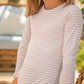 A child in a long-sleeve Minnow Girls' Rashguard One Piece swimsuit with pink checkered pattern, grosgrain ribbon accents, and ruffled edges sits outdoors, enjoying the sun while staying sun-safe.